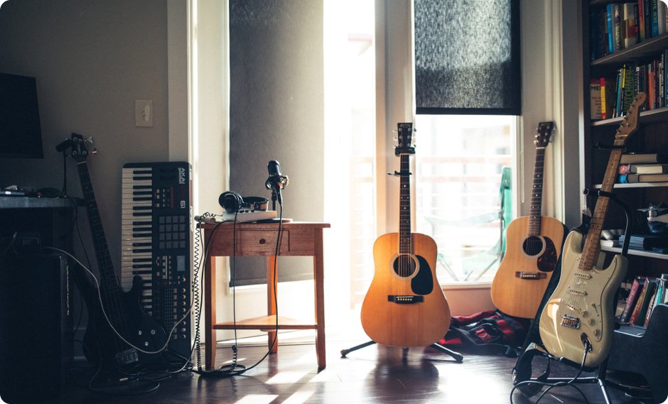 In einem Raum mit Bücherregal stehen 4 Gitarren, ein an die Wand gelehntes Keyboard und auf einem Tisch Musikequipment